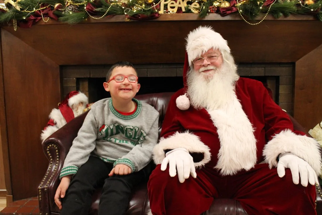 A young boy sitting next to Santa Claus on a decorated Christmas mantel, smiling, with festive decorations in the background.