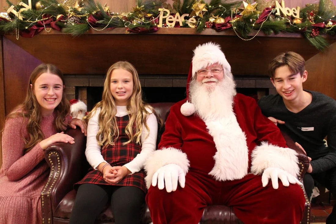 Three children and Santa Claus sitting on a leather couch in front of a decorated fireplace with a festive garland, holidays signs, and ornaments.