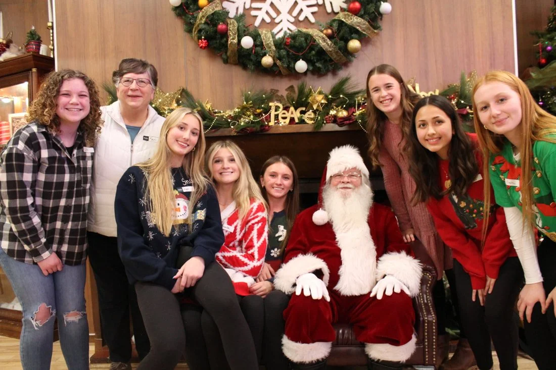 Group of nine people, including children and adults, gathered around Santa Claus at a Christmas celebration, with holiday decorations and a wreath in the background.