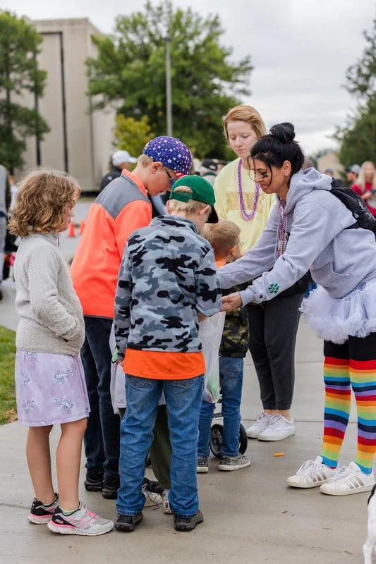 Kids and adults participating in an outdoor event, some children are receiving or exchanging items or prizes. The setting is on a sidewalk with trees and a building in the background.