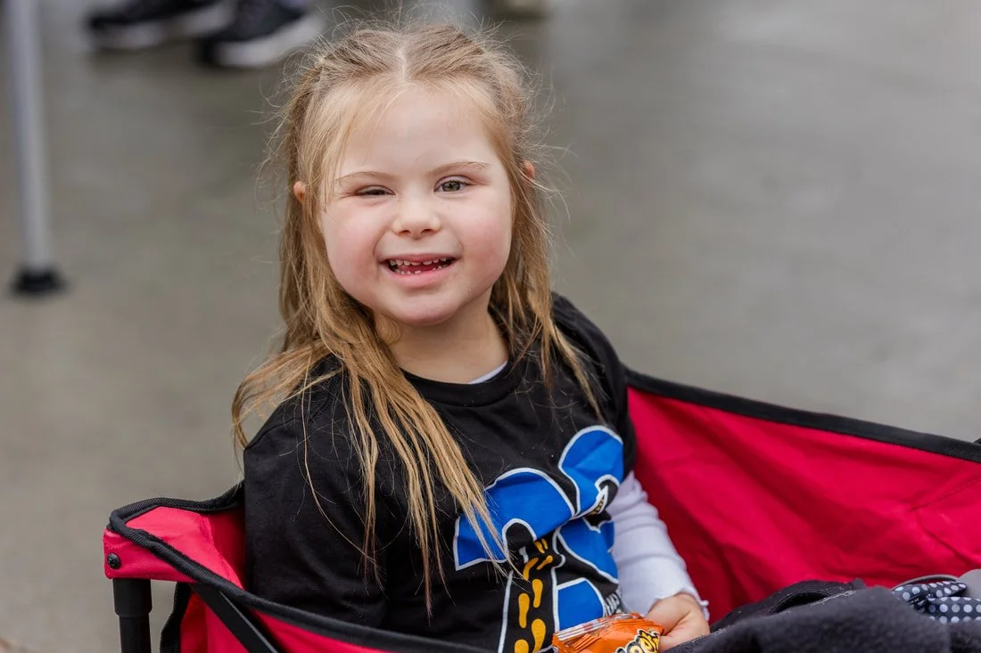 A young girl with long, light brown hair, smiling with missing front teeth, sitting in a red and black stroller outdoors, holding a bag of snacks.