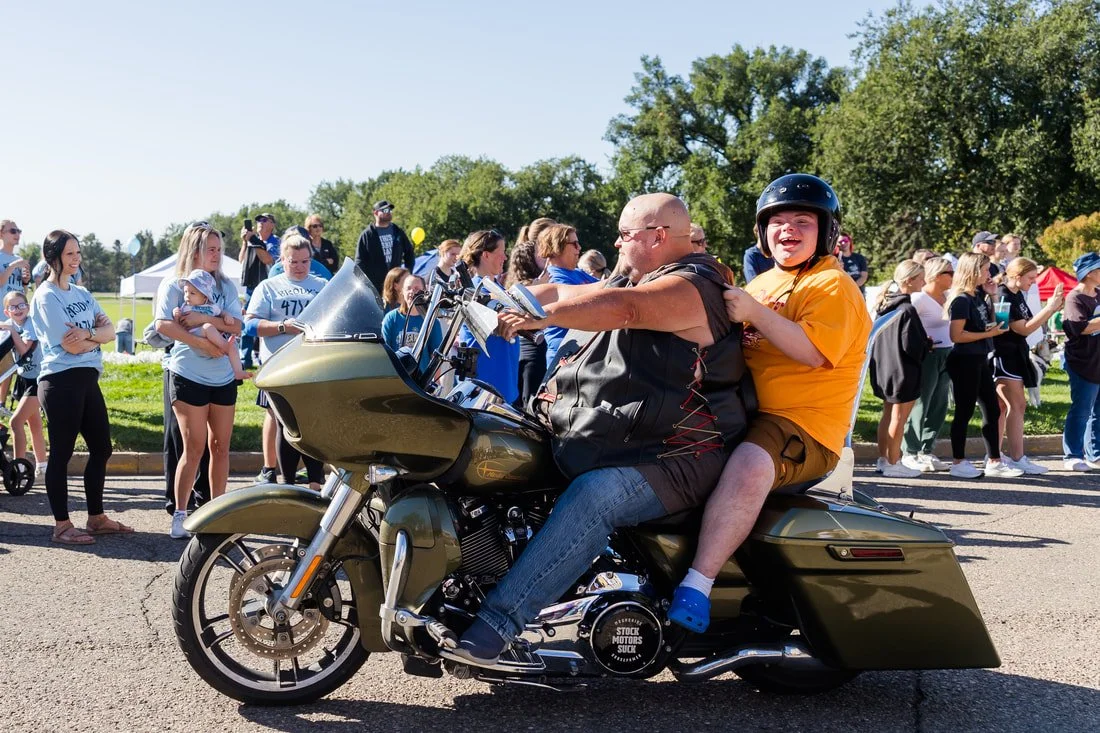 A man and a boy riding a motorcycle at an outdoor event with many onlookers, including women and children, on a sunny day.