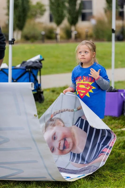 A young girl with blonde hair, wearing a blue Superman t-shirt, stands outdoors on a grassy area near a park. She is holding a large banner with the face of a smiling child printed on it, and there are tents and trees in the background.