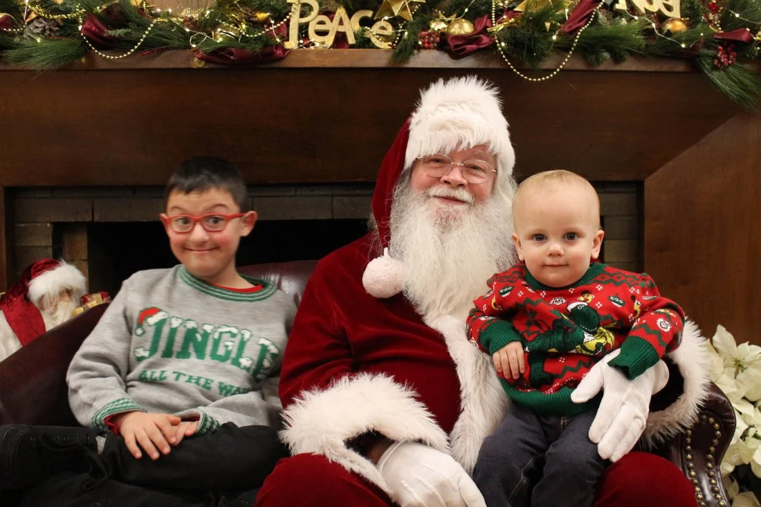 Santa Claus sitting on a couch with two children, a boy wearing glasses and a girl in an elf sweater, in front of a Christmas fireplace decorated with greenery, gold ornaments, and a 'Peace' sign.