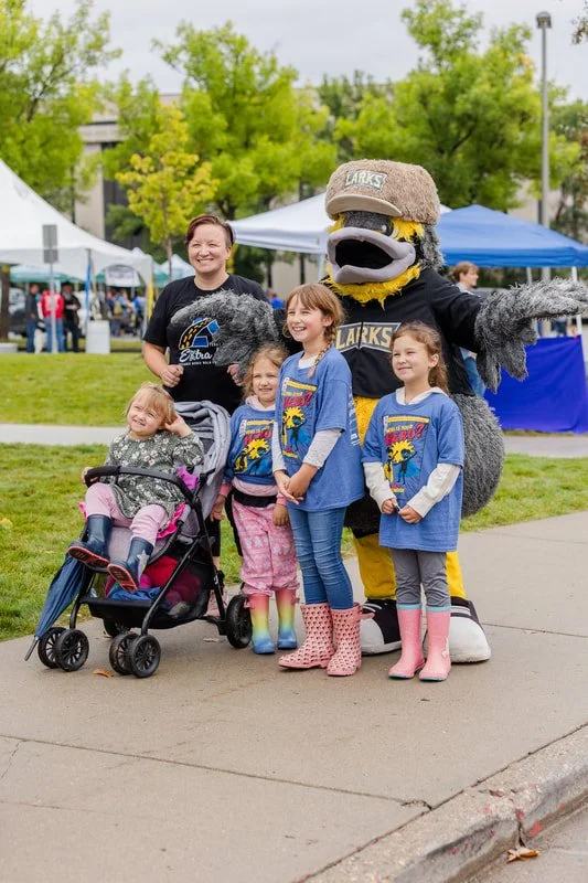 A group of children and a woman standing outdoors with a mascot dressed as a bird, at a community event under tents and trees.