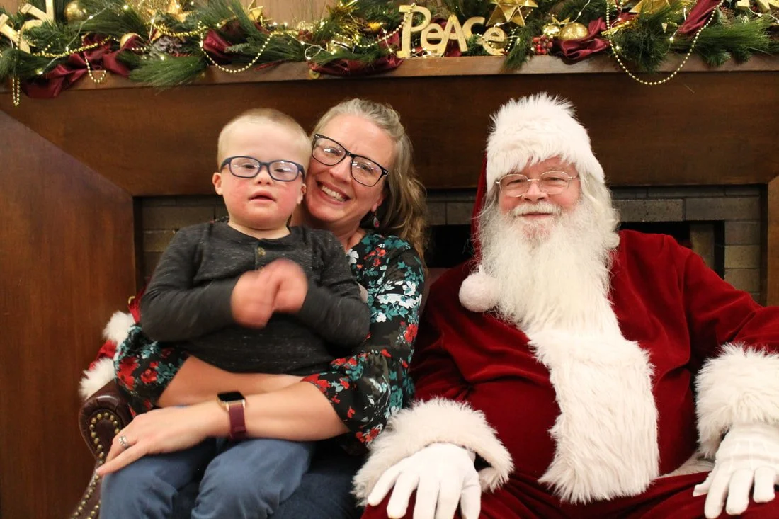 A woman and a young boy with glasses sitting with Santa Claus in front of a Christmas fireplace.