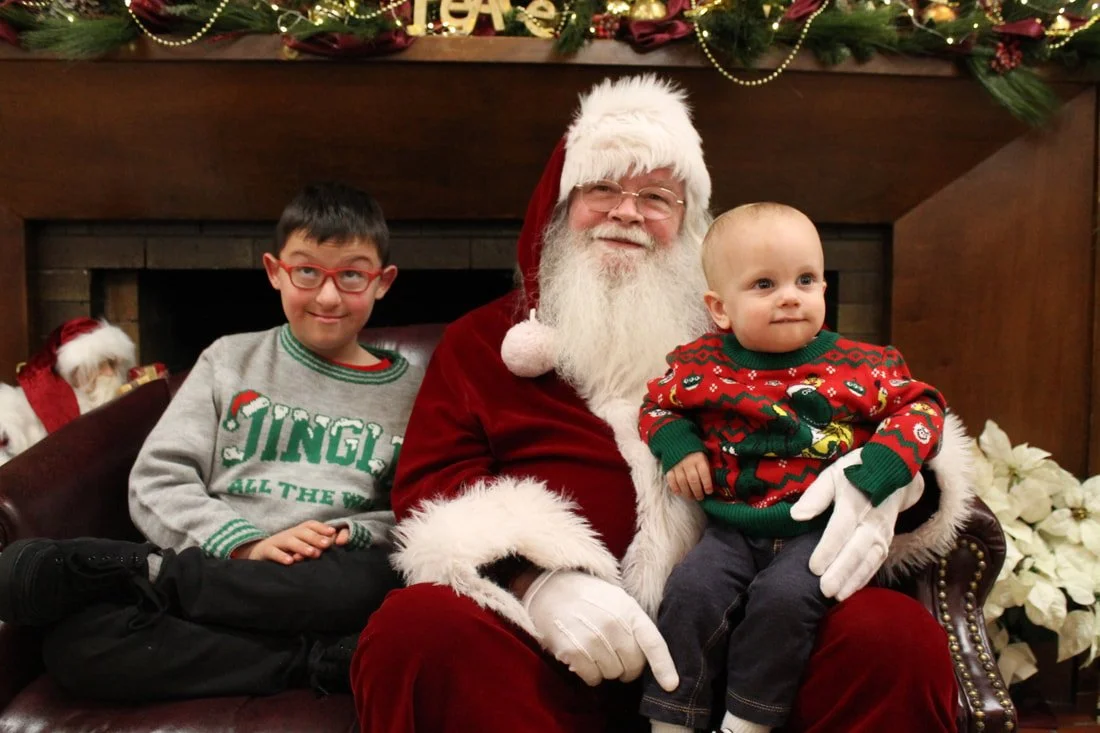 Two children sitting with Santa Claus in a Christmas setting, decorated with festive garlands and poinsettias.