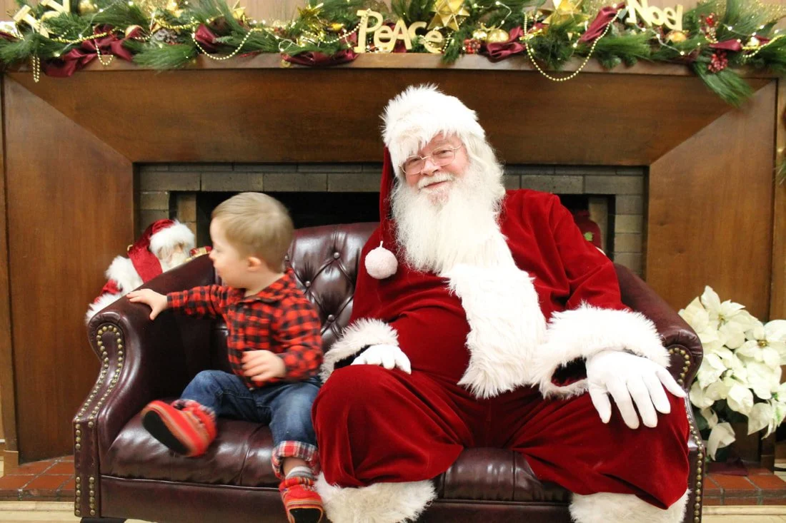 A child sitting next to Santa Claus on a leather sofa in front of a fireplace decorated with Christmas ornaments and greenery.