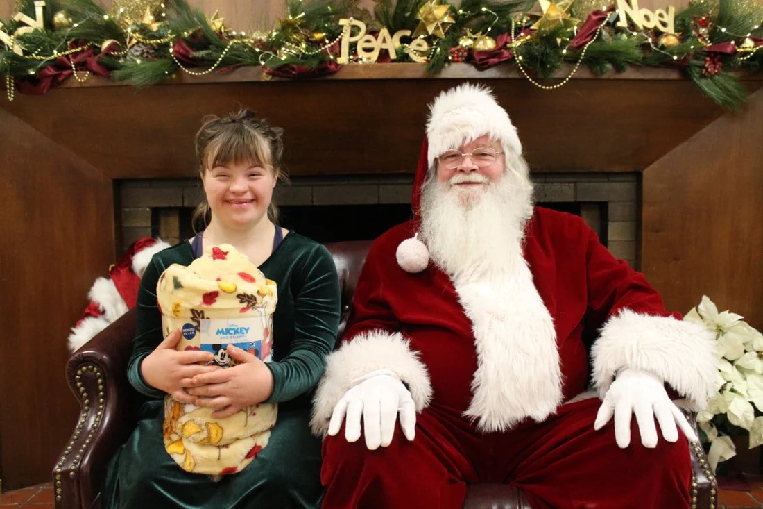 A girl in a green dress sitting next to Santa Claus in a festive decorated room. The girl is holding a wrapped gift, and both are smiling. Christmas decorations, including garlands, ornaments, and a fireplace, are visible in the background.