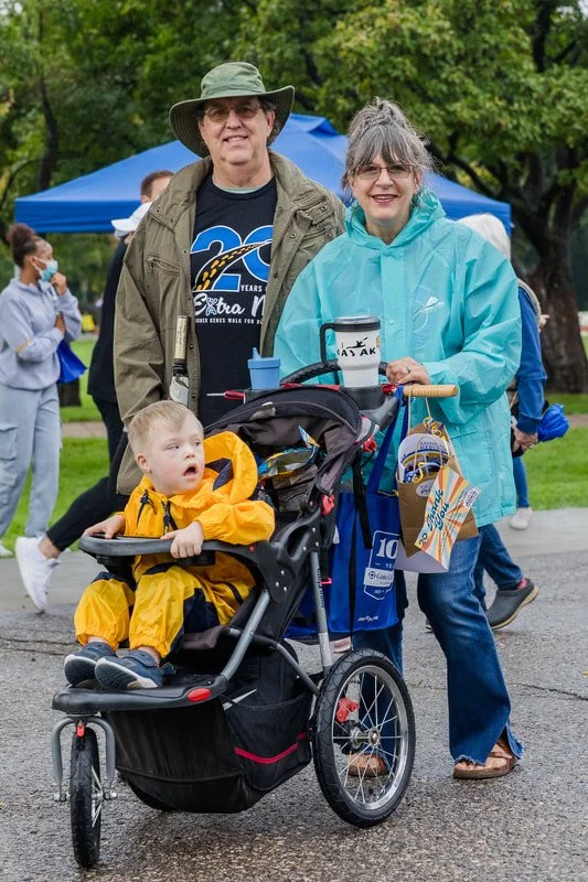 An older couple standing outdoors with a young child in a stroller, at an event under a blue canopy. The woman is wearing a light blue jacket, and the man has a gray hat and glasses. The child is dressed in a yellow raincoat, looking surprised or cur