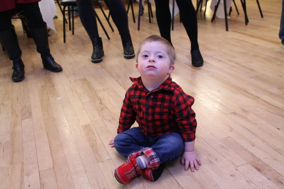 A young boy sitting on a wooden floor, looking up at the camera, with people standing behind him.