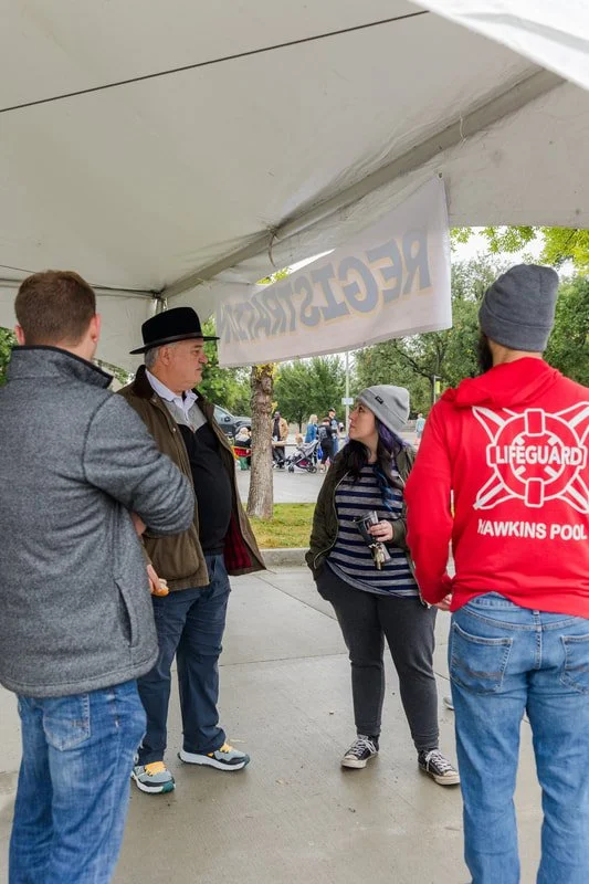 Four people standing under a tent, engaged in conversation, with trees and a park setting in the background. One person wears a lifeguard hoodie, while another wears a fedora and a jacket.