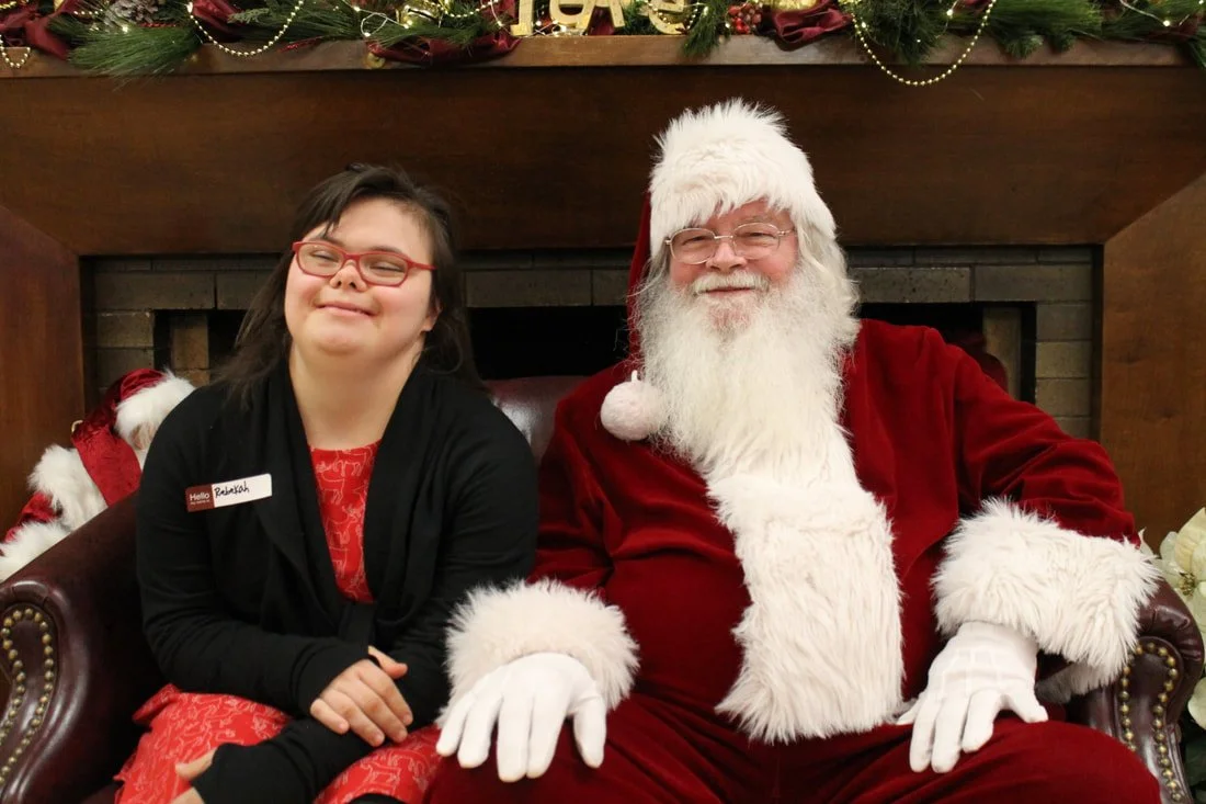 A young woman with glasses sitting next to a man dressed as Santa Claus in front of a decorated fireplace