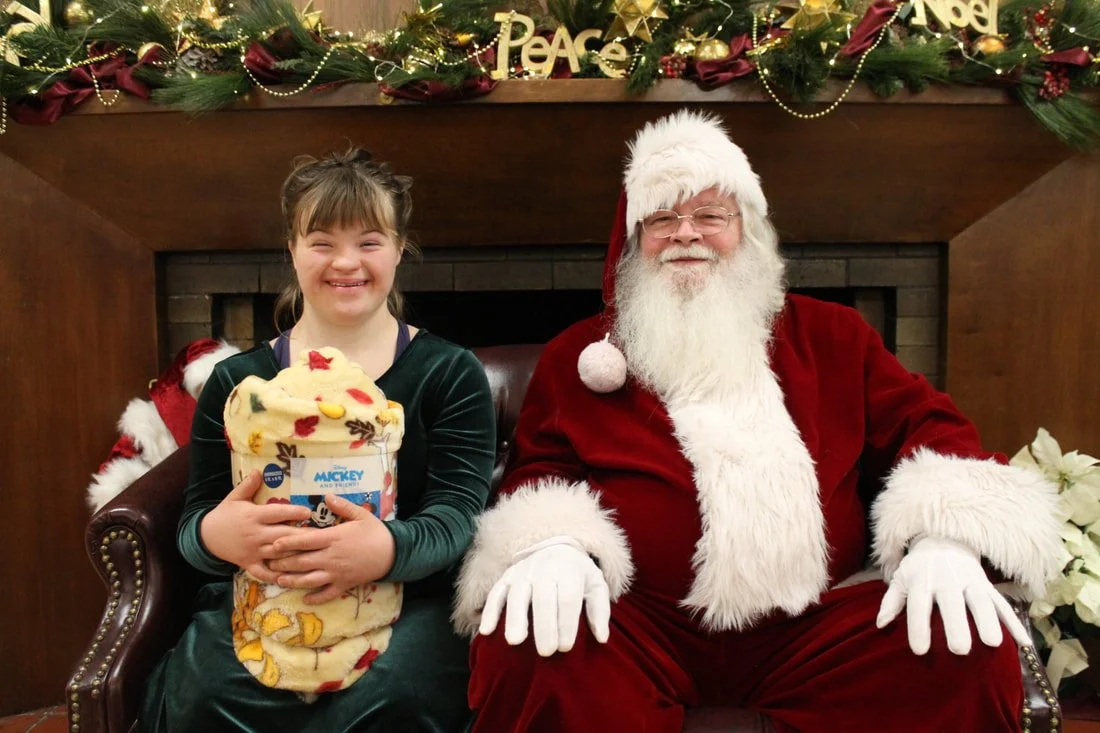 A young girl sitting next to Santa Claus, both smiling at the camera during a Christmas celebration. The girl is holding a wrapped present. The background is decorated with Christmas garland, ornaments, and the words "Peace" and "Noel."