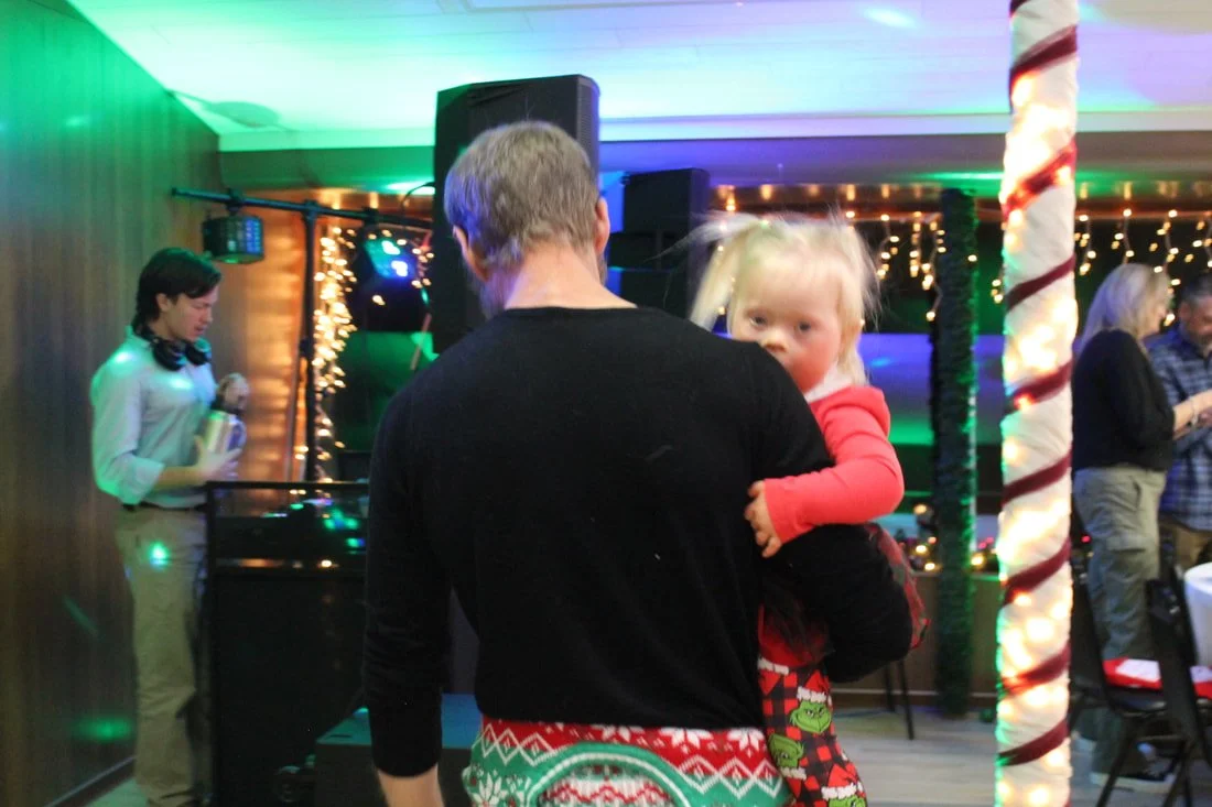 A man holding a young girl in a decorated room with Christmas lights and holiday decorations, including a large candy cane striped pole, at a Christmas party.