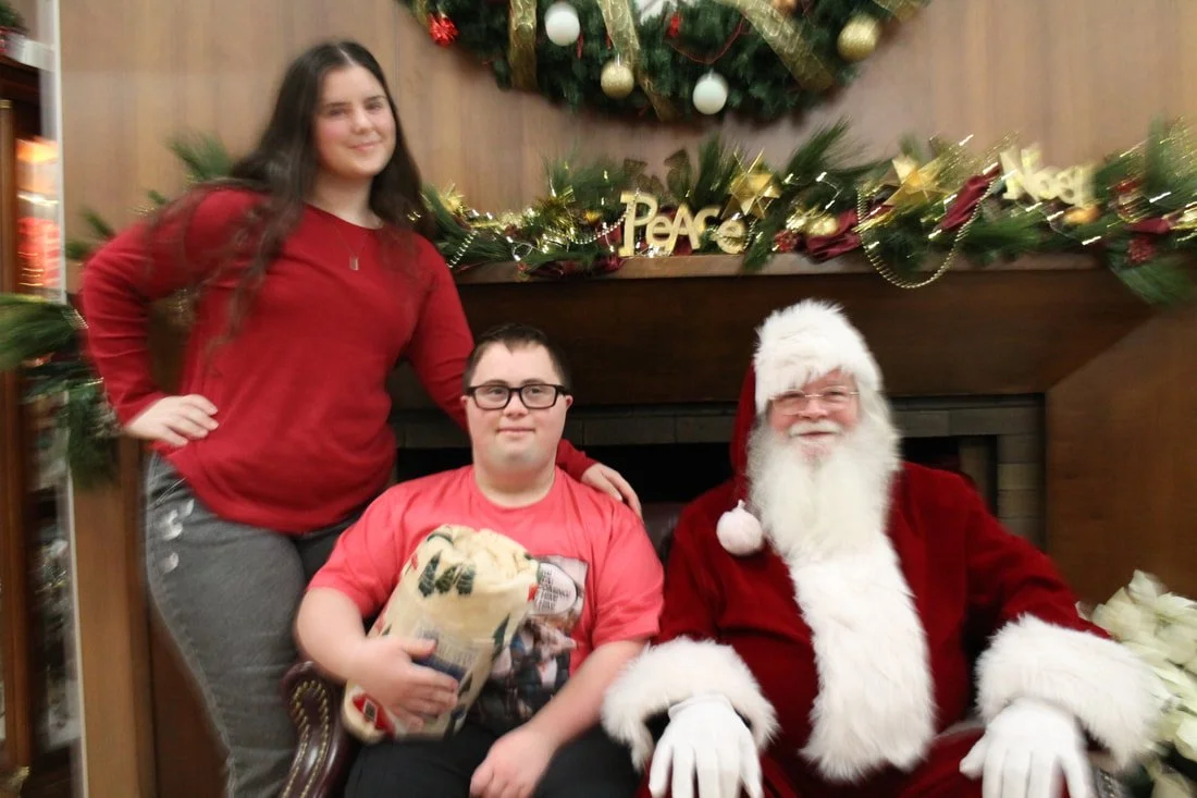 A girl and a boy sitting with Santa Claus in front of a decorated fireplace with a Christmas wreath, ornaments, and a sign that says 'Peace'.
