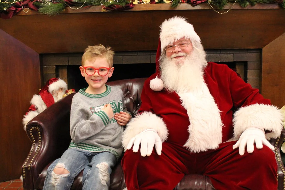 A young boy with red glasses sitting next to Santa Claus, who is wearing a red suit with white fur trim, in front of a fireplace decorated with Christmas garland.