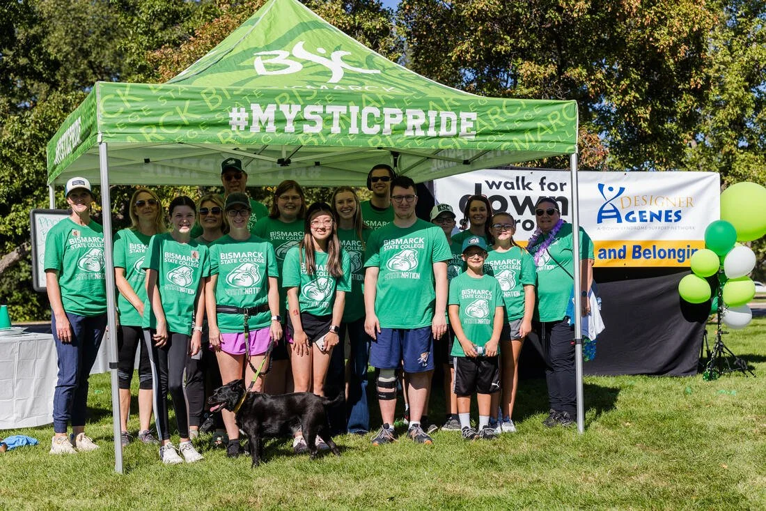 Group of people, mostly children, standing under a bright green canopy with hashtag #MYSTICPRIDE. They wear matching green T-shirts with Bismarck State College logo, and there are colorful balloons on the side. The background is a park with trees and