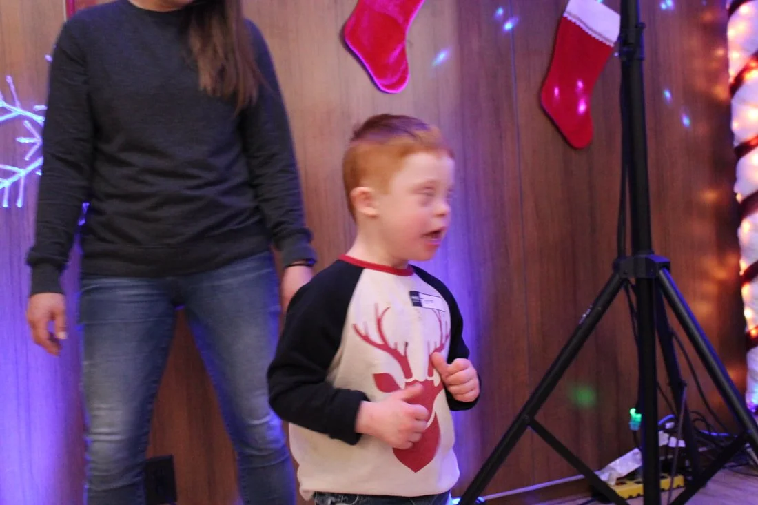 A young boy with red hair, wearing a Christmas-themed shirt with a reindeer design, is standing in a festive setting with Christmas stockings and colorful lights on a wooden wall. A woman in a dark sweater and jeans stands nearby.