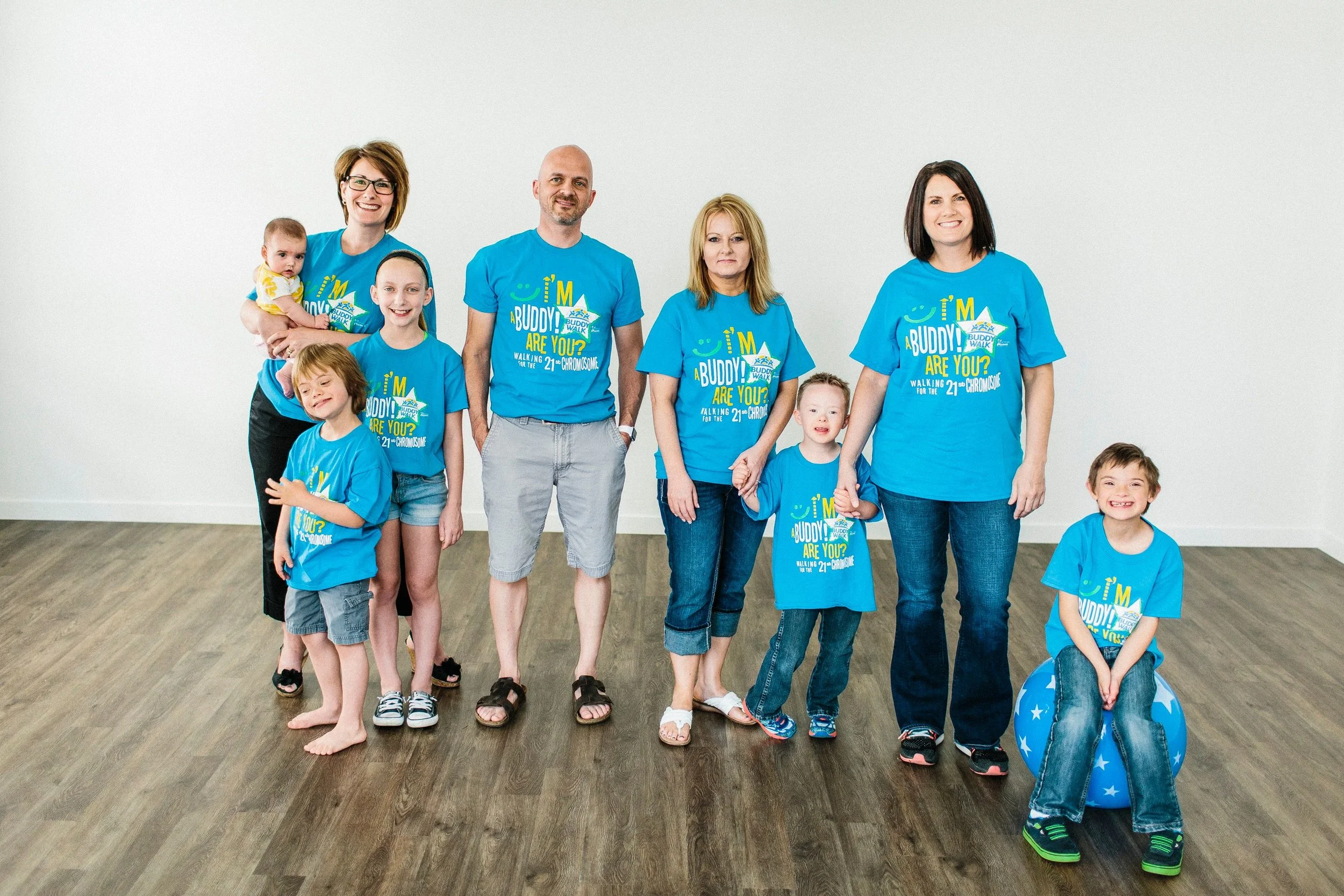 Group of adults and children wearing matching blue t-shirts with event branding, standing on a wooden floor inside a plain room.