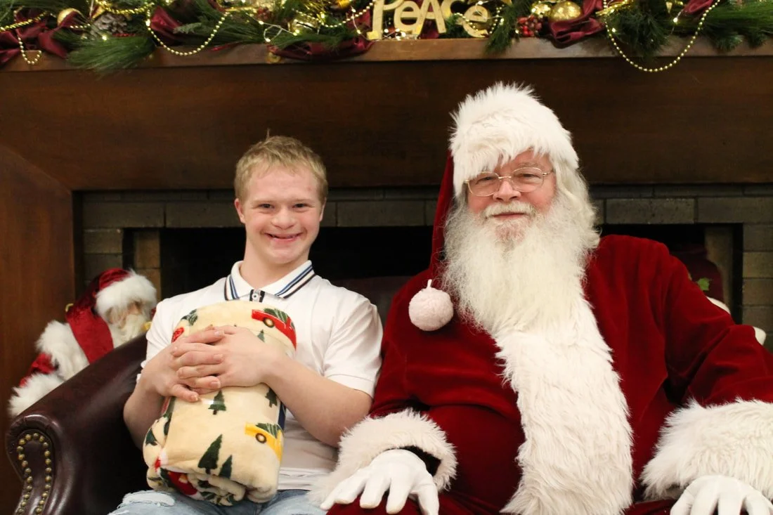 A young boy sitting next to Santa Claus in front of a decorated fireplace with Christmas ornaments. The boy is holding a wrapped gift and smiling. Santa is dressed in a red suit with white fur trim, wearing glasses and a Santa hat, also smiling.