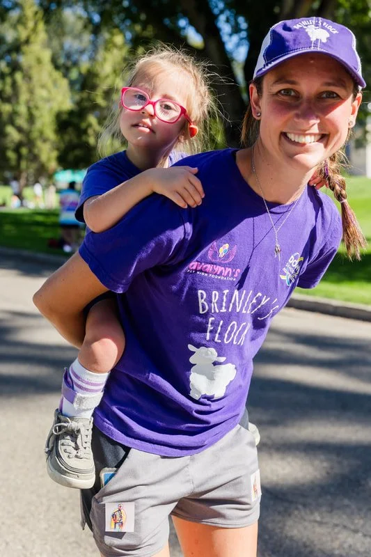 A woman with a young girl on her back, both smiling, in a park on a sunny day.