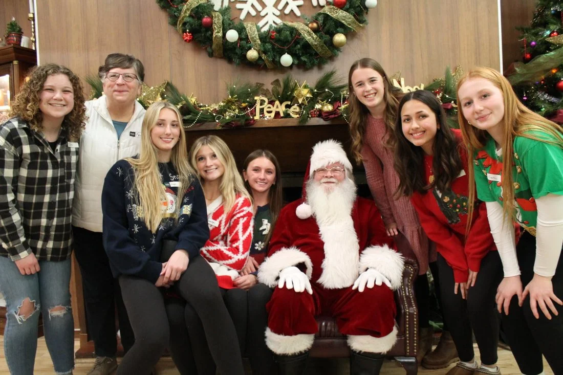 Group of children and adults gathering around Santa Claus seated on a decorated chair in front of a Christmas wreath and holiday decorations.