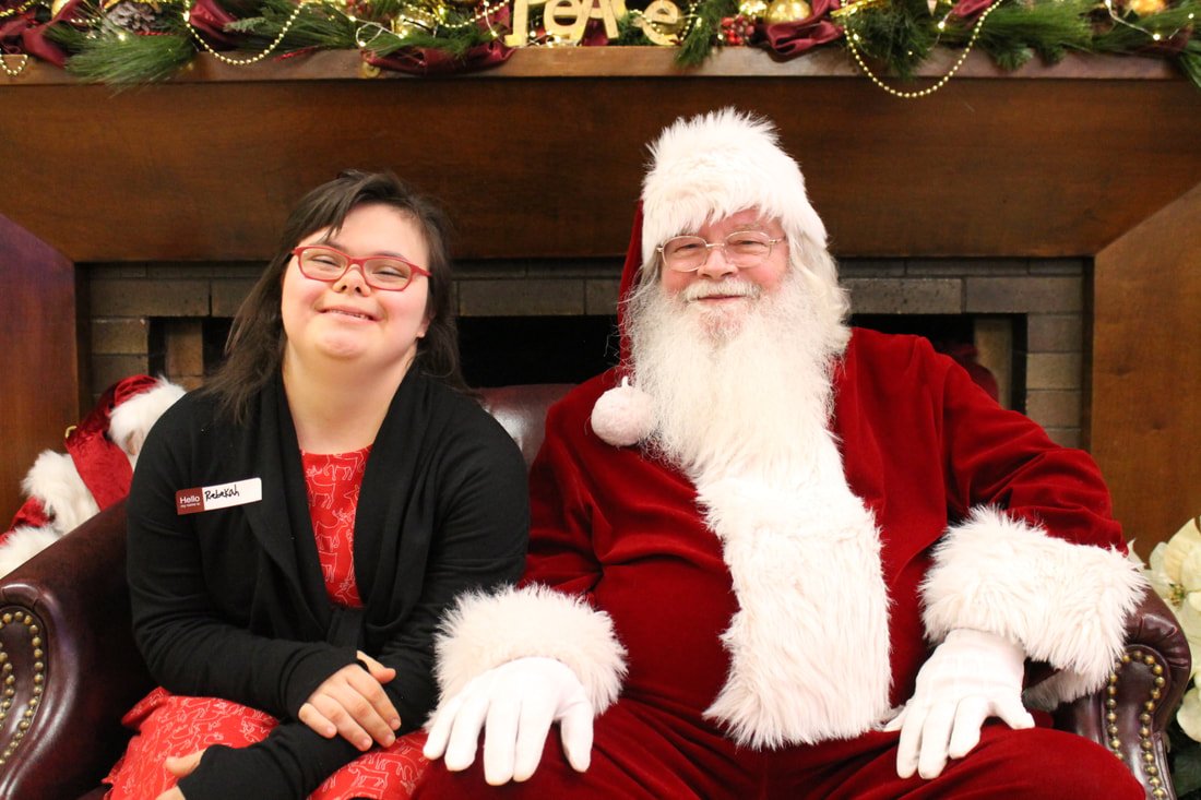 A young girl wearing glasses and a red dress sitting next to a man dressed as Santa Claus on a couch in front of a decorated fireplace with Christmas garland and lights.