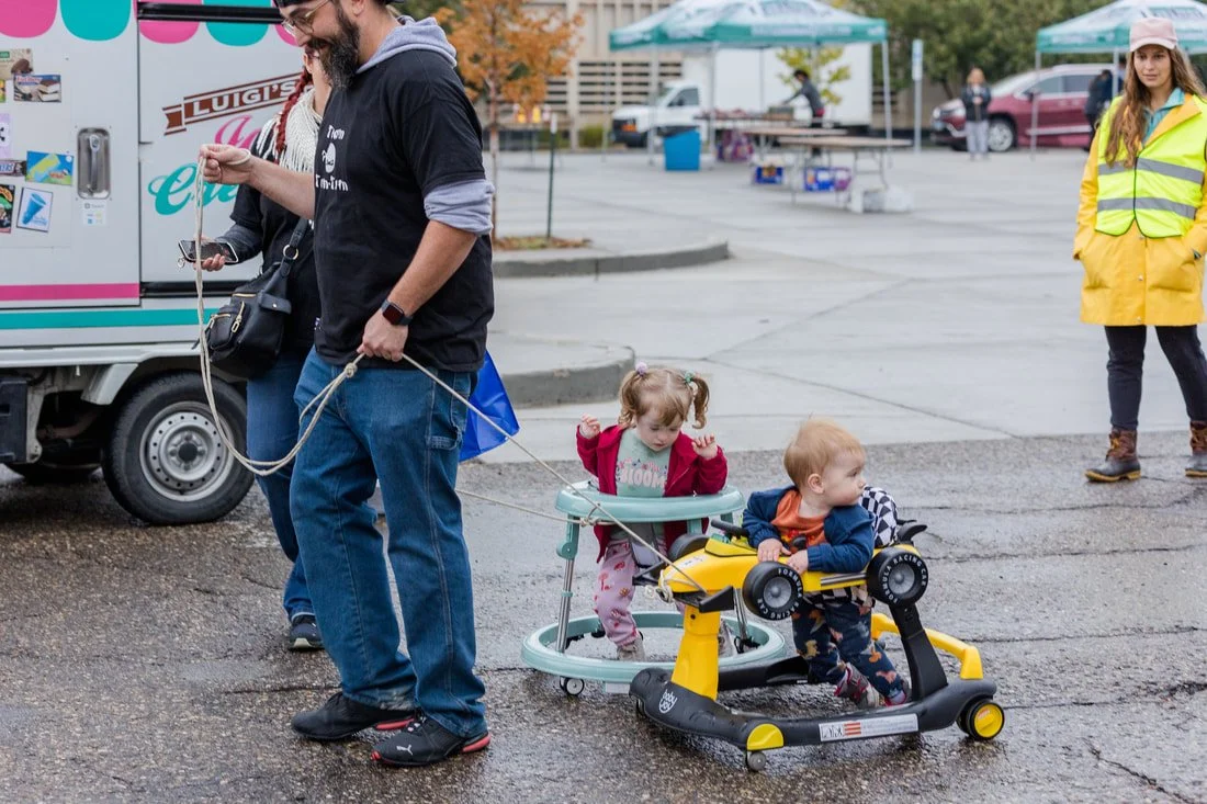 A man holding a leash attached to two young children sitting in a toy car and a stroller in a parking lot, with a woman in a yellow safety vest observing nearby.