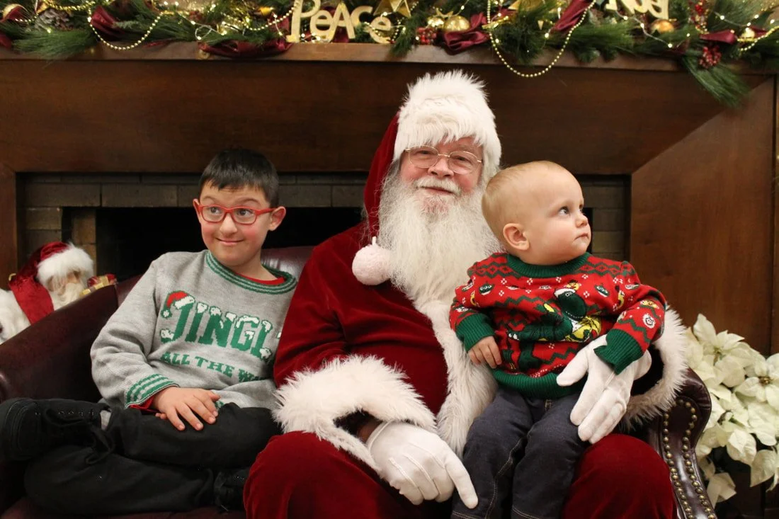 A festive Christmas scene with Santa Claus sitting on a chair, holding a young child dressed in a holiday sweater, while an older boy in glasses sits beside him. The background is decorated with Christmas garlands, ornaments, and poinsettias.