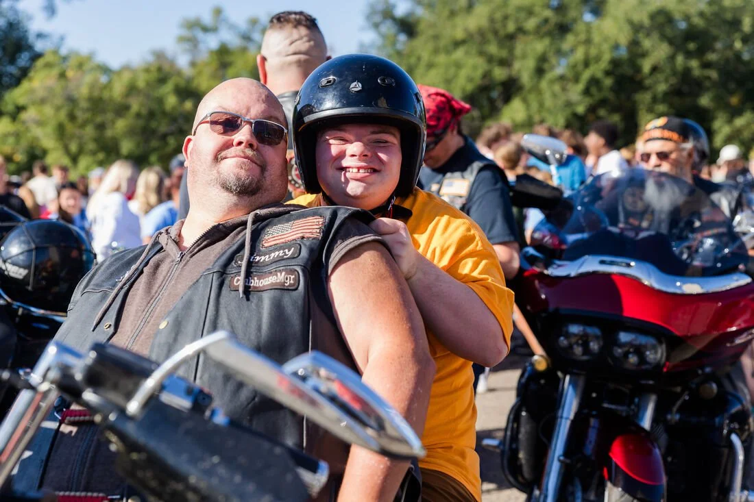 A man and a boy smiling, with the boy wearing a helmet, surrounded by motorcycles and other people outdoors.