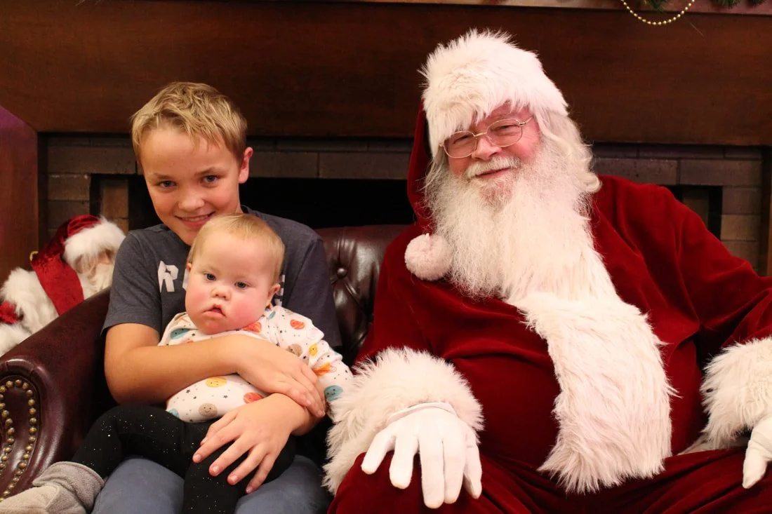 A young boy and a baby sitting with a man dressed as Santa Claus in a festive setting.