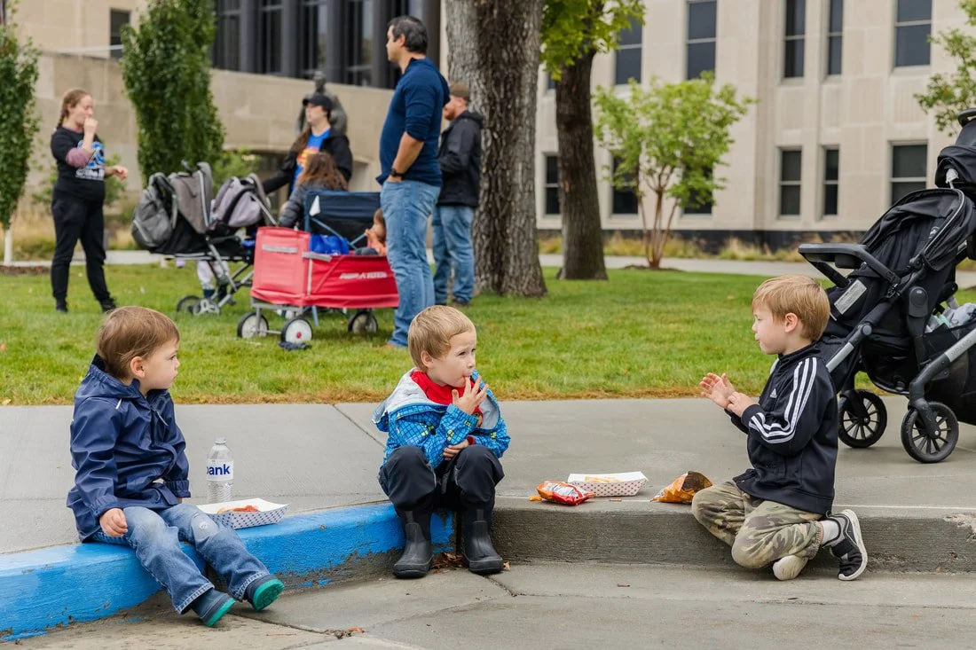 Three children sitting on a curb enjoying snacks, with adults and strollers in the background on a city sidewalk.
