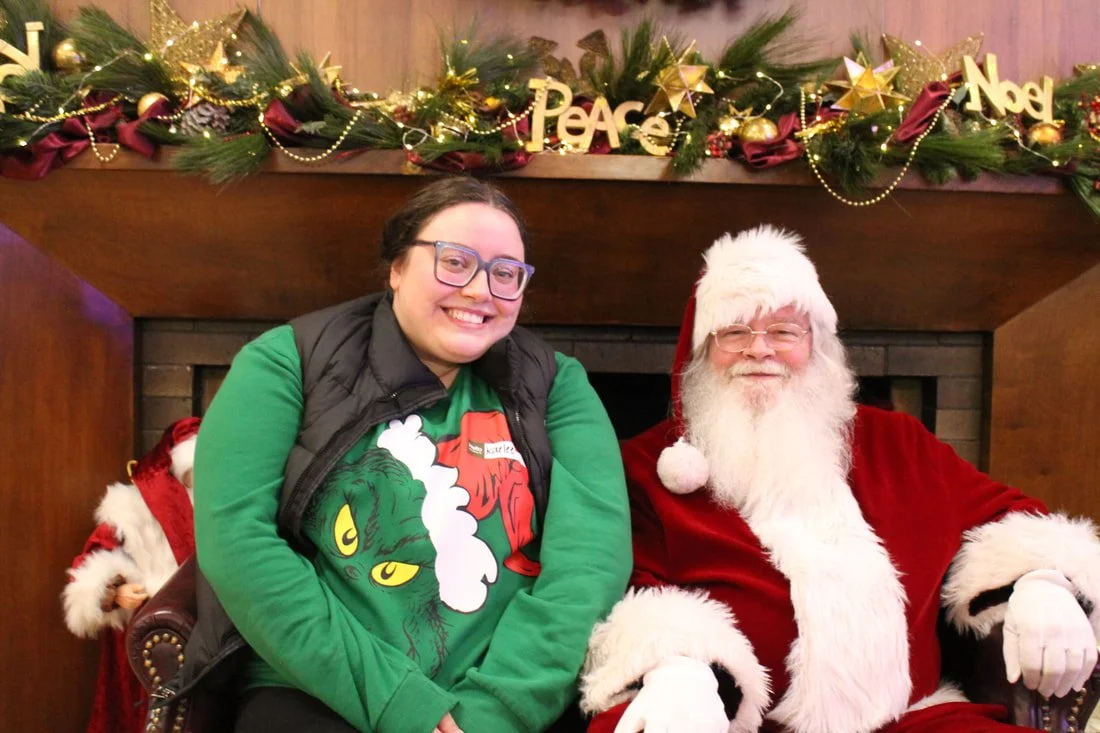 A woman and Santa Claus sitting together by a decorated fireplace, smiling for the camera. The woman is wearing a green Christmas sweater featuring the Grinch, and Santa is dressed in his traditional red suit with white trim. The fireplace mantel is 