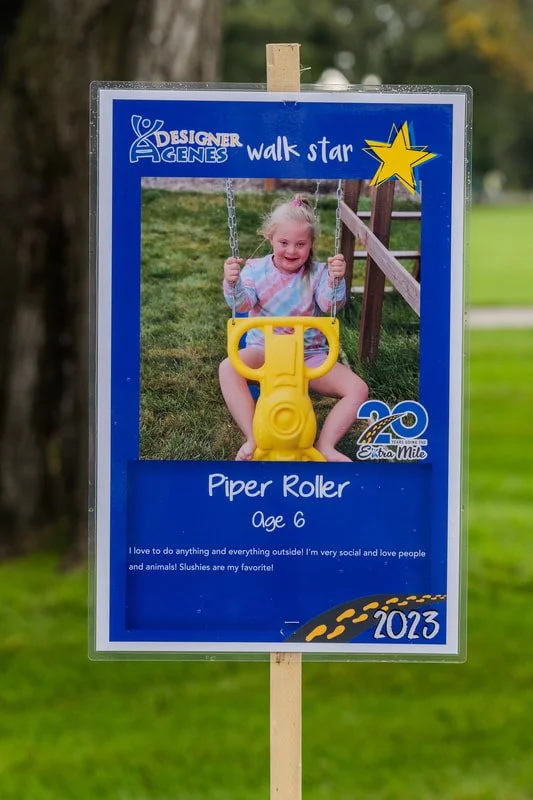 A sign featuring a young girl named Piper Roller, age 6, on a playground swing with a yellow plastic seat, smiling. The sign includes her favorite activities, the year 2023, and is part of the Designer Genes Walk Star event.