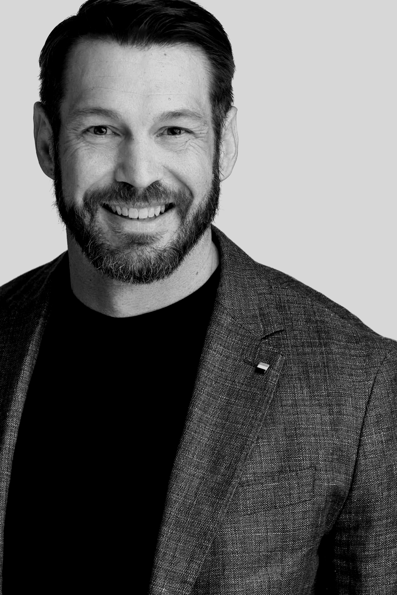 A smiling man with a beard and mustache, wearing a dark shirt and a textured blazer, against a plain background.