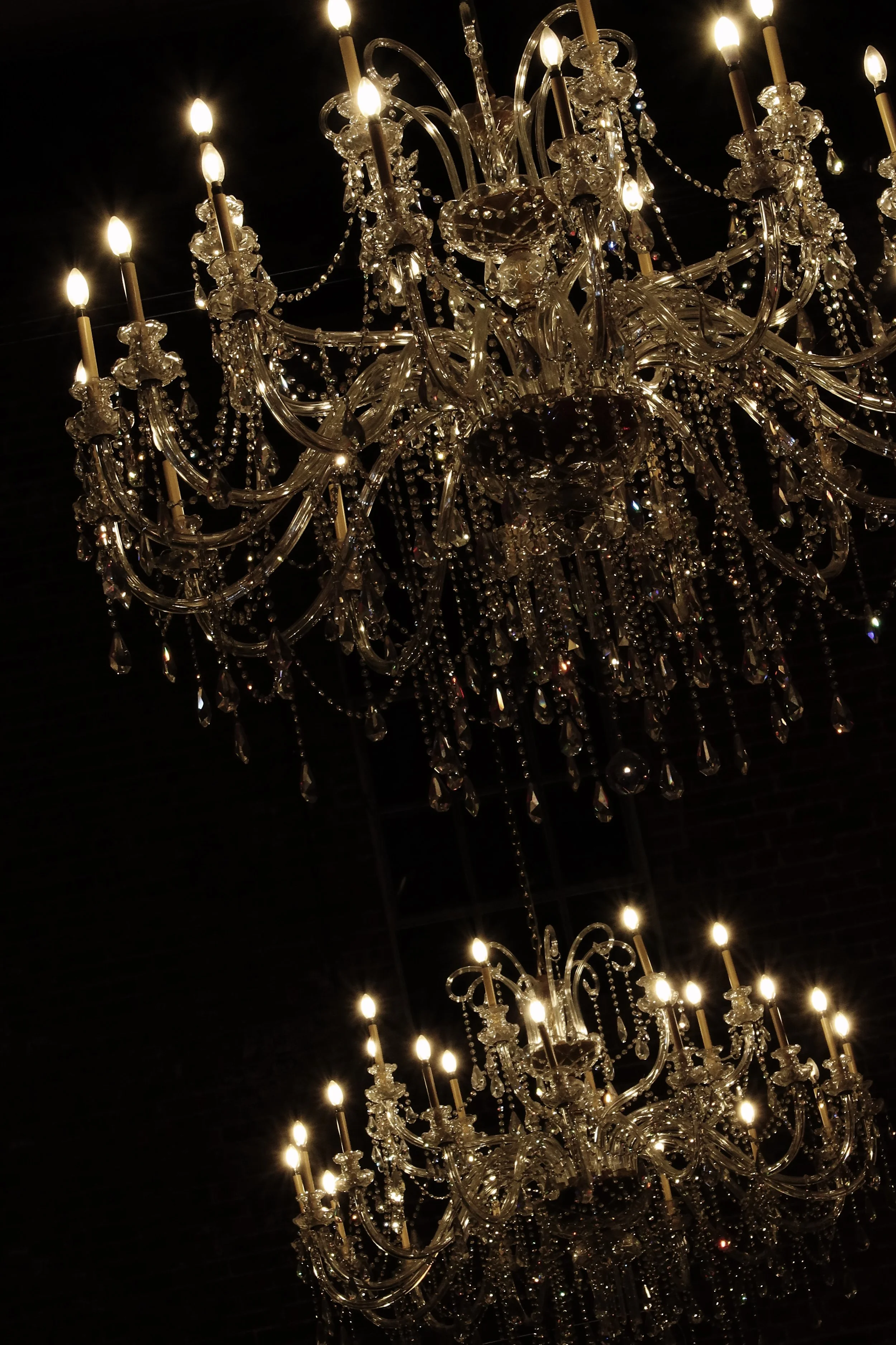 Two ornate crystal chandeliers with lit candle-shaped lights hanging from a dark ceiling.