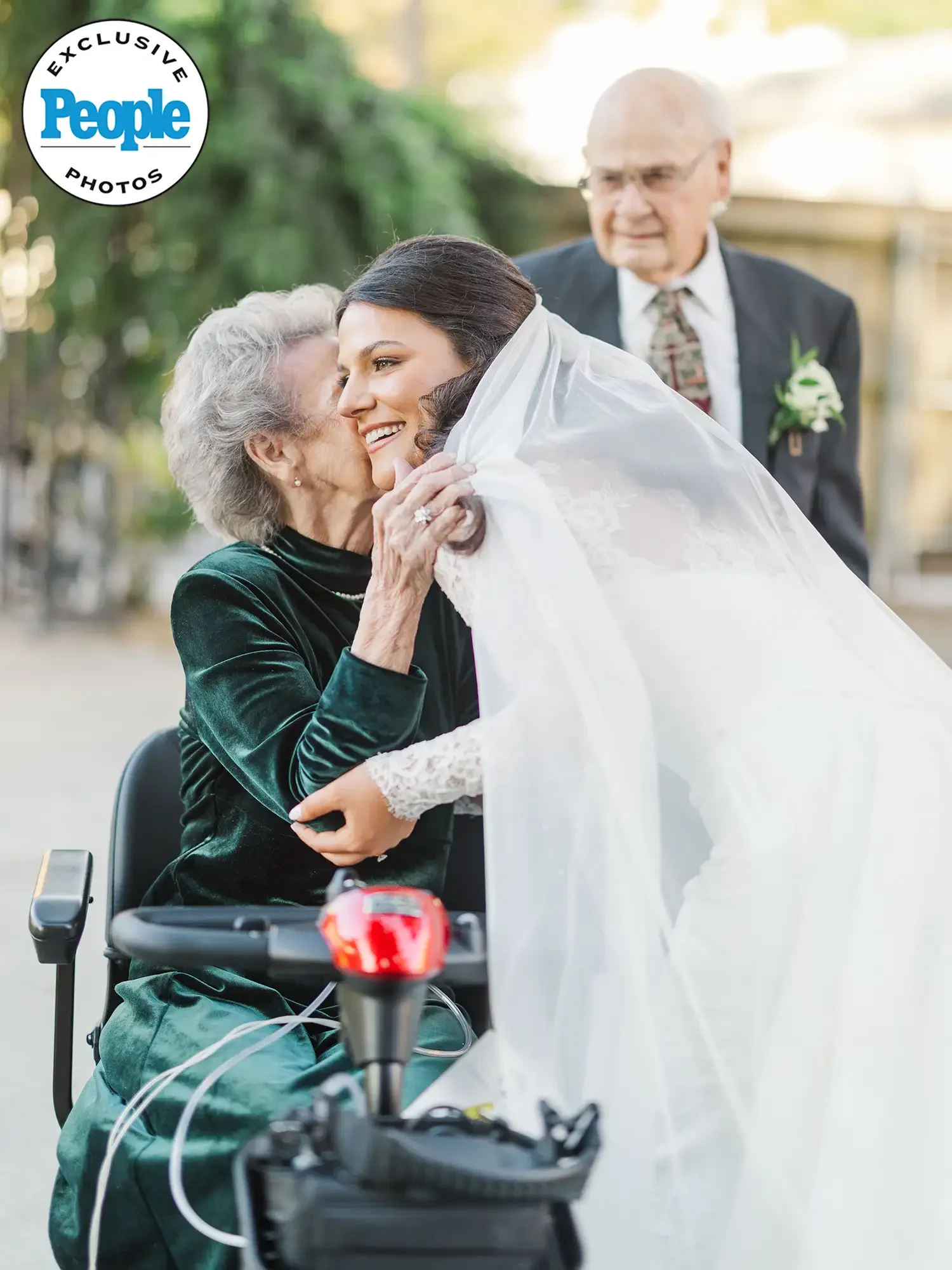 An elderly woman in a wheelchair is hugging a young woman in a wedding dress while a man in a suit looks on in the background outdoors.
