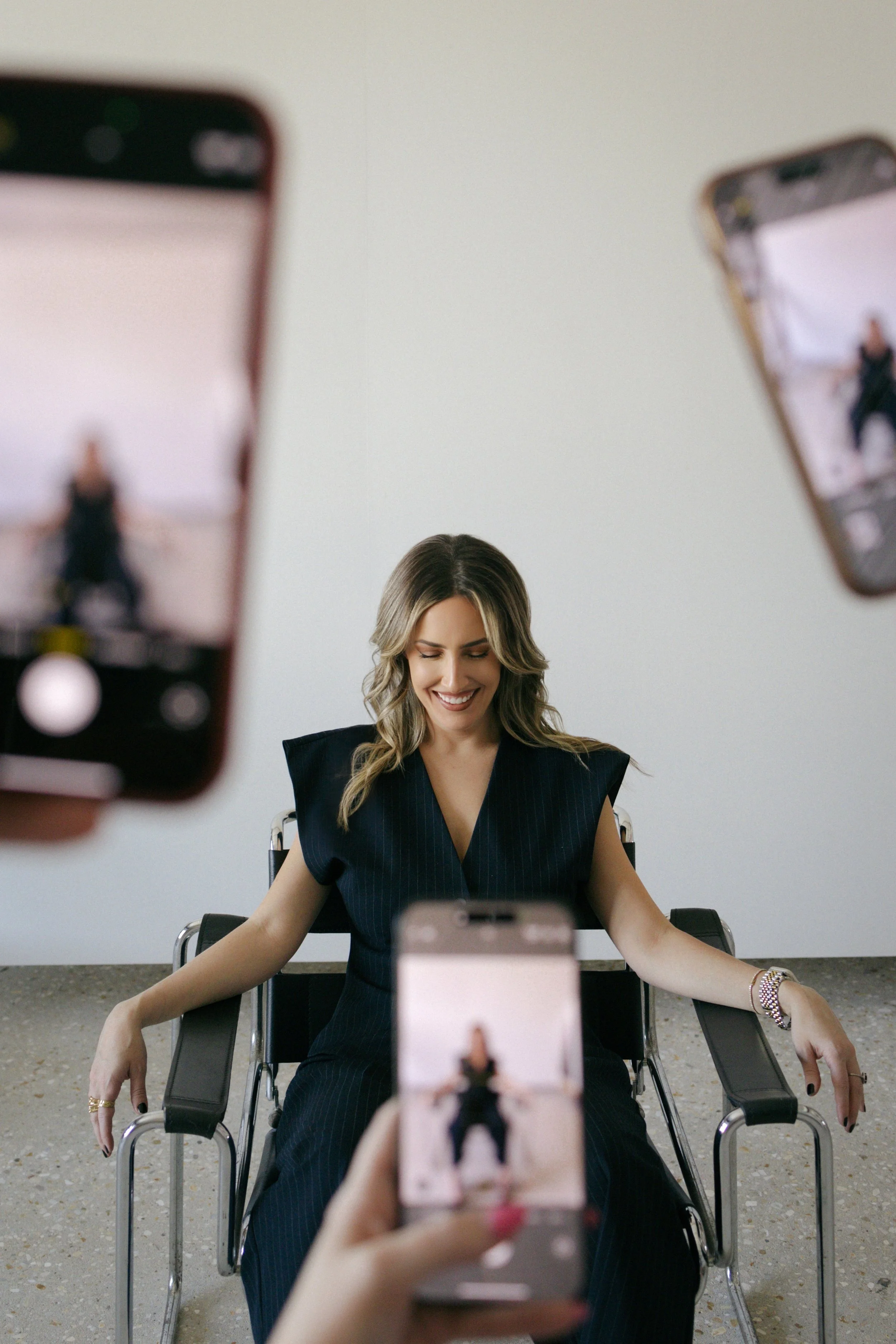 A woman sitting in a chair smiling as she is photographed by three people. The photo is being taken by three smartphones held in front of her.
