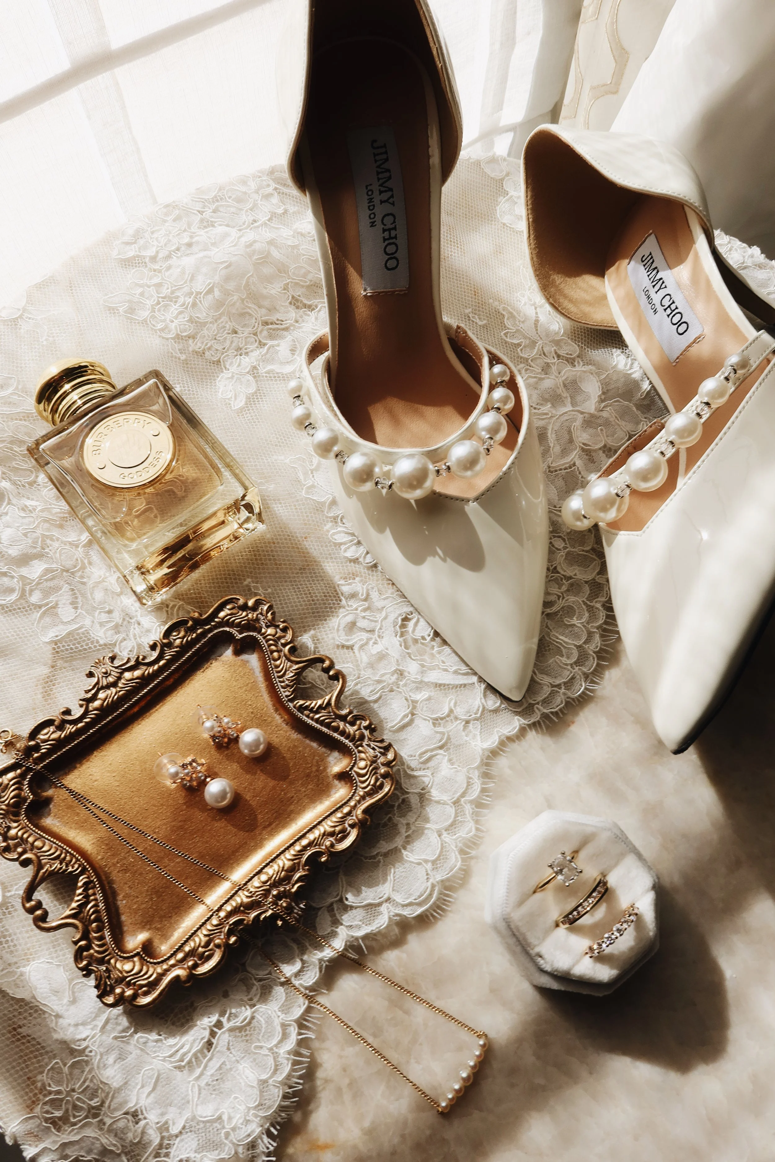 A pair of cream-colored Jimmy Choo high heels decorated with large pearl embellishments, placed on a lace doily. Nearby are a bottle of perfume, gold jewelry including earrings and rings, and a necklace displayed on a decorative gold tray, all on a lace-covered surface.