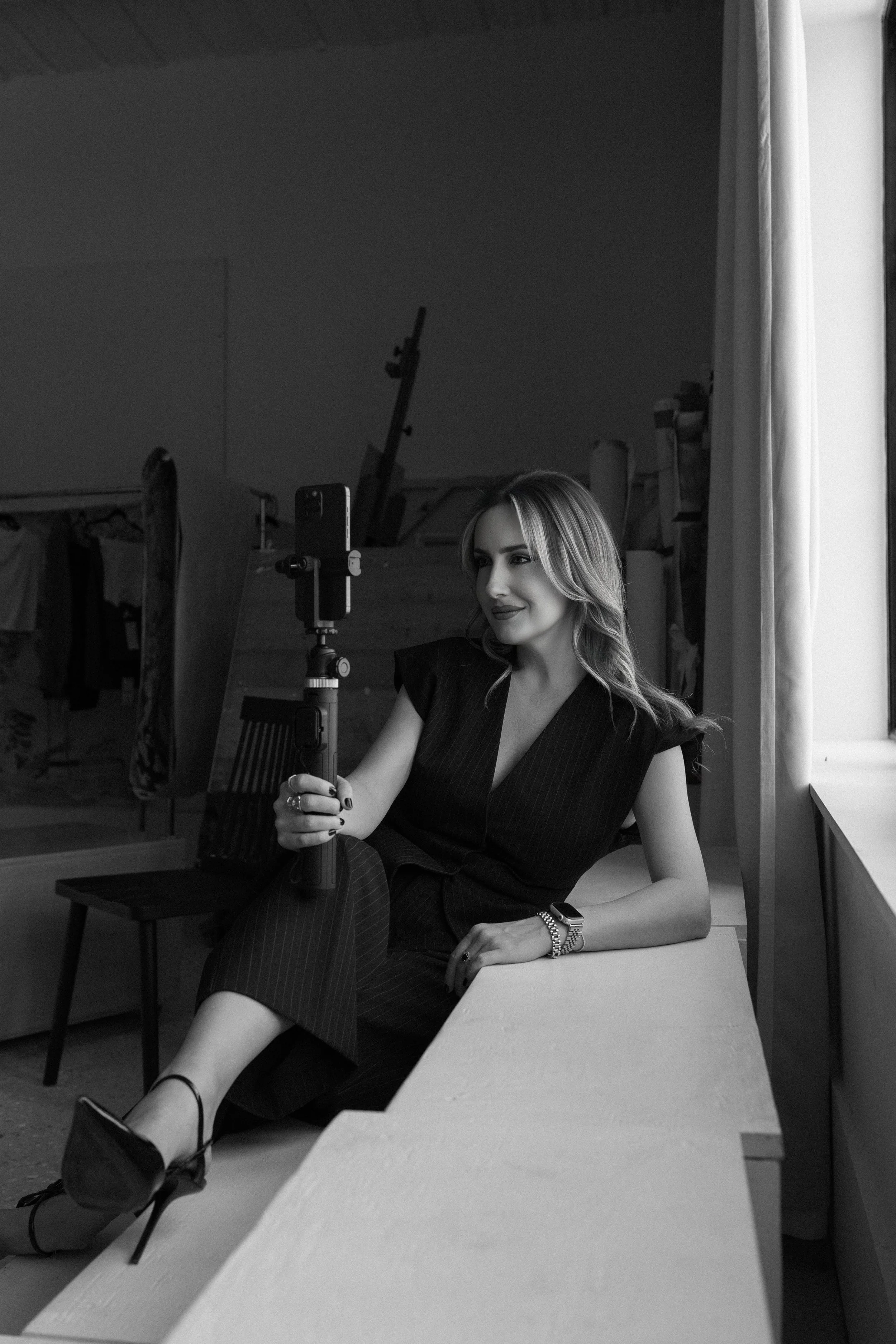 A woman in a black pinstripe dress sitting on a bench by a window, taking a selfie with her smartphone mounted on a selfie stick in a room with draped fabrics and studio equipment.