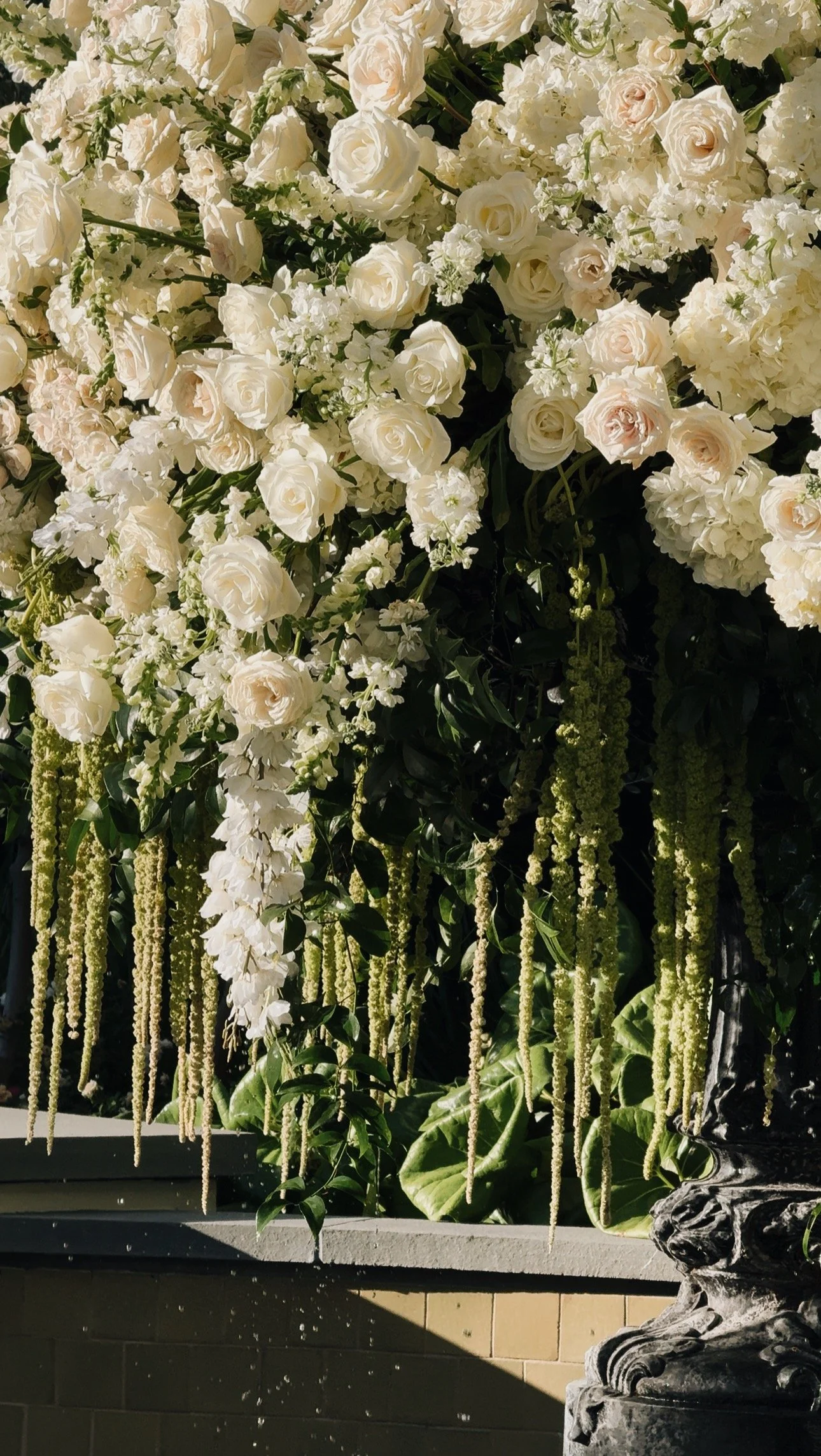 A floral arrangement featuring white roses, white hydrangeas, and hanging white and green floral strands, set in a black ornate vase on a ledge.