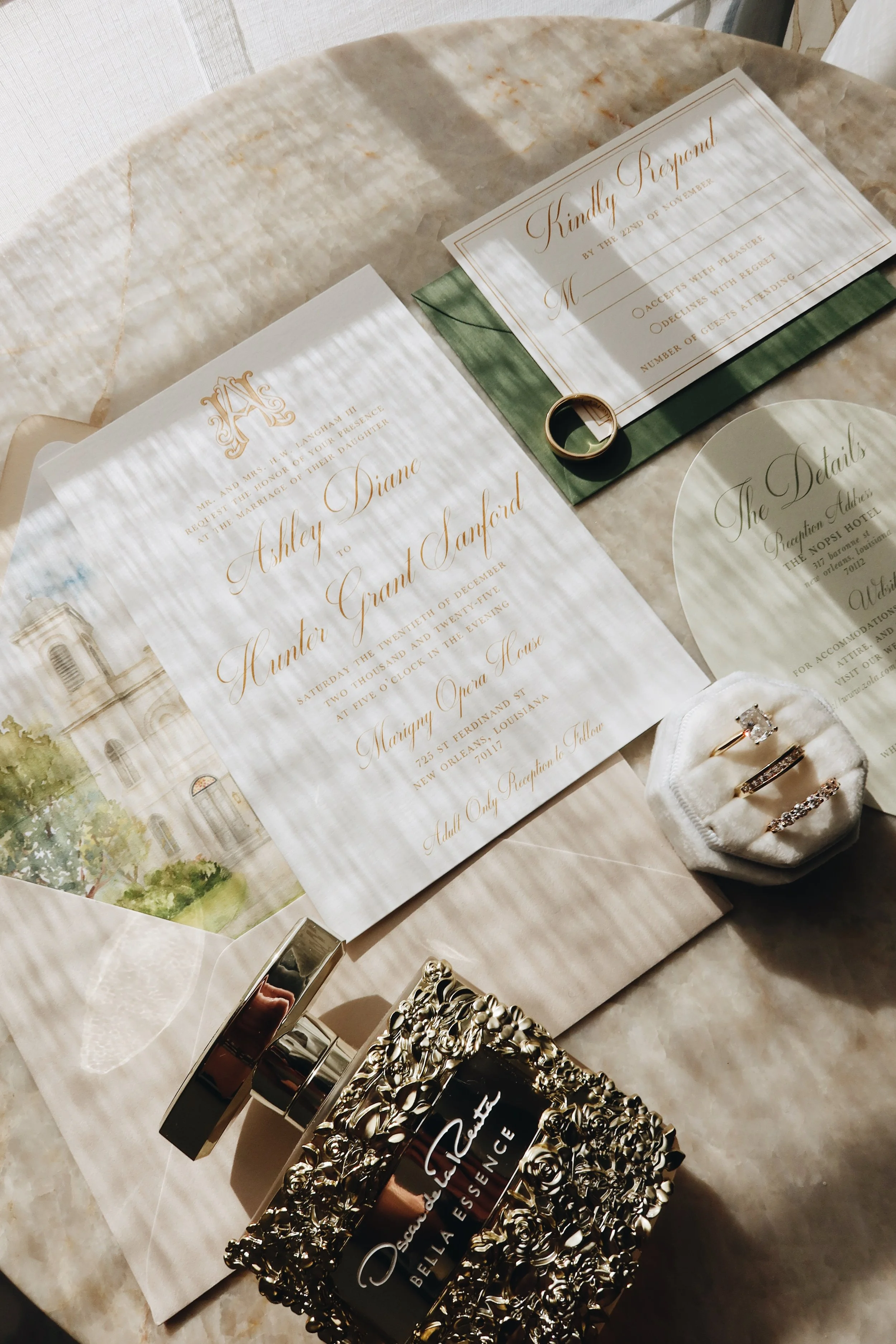 Wedding invitation, rings, and perfume on a marble table. The invitation is for Ashley Diane and Hunter Grant Sanford's wedding at Marigny Opera House in New Orleans.