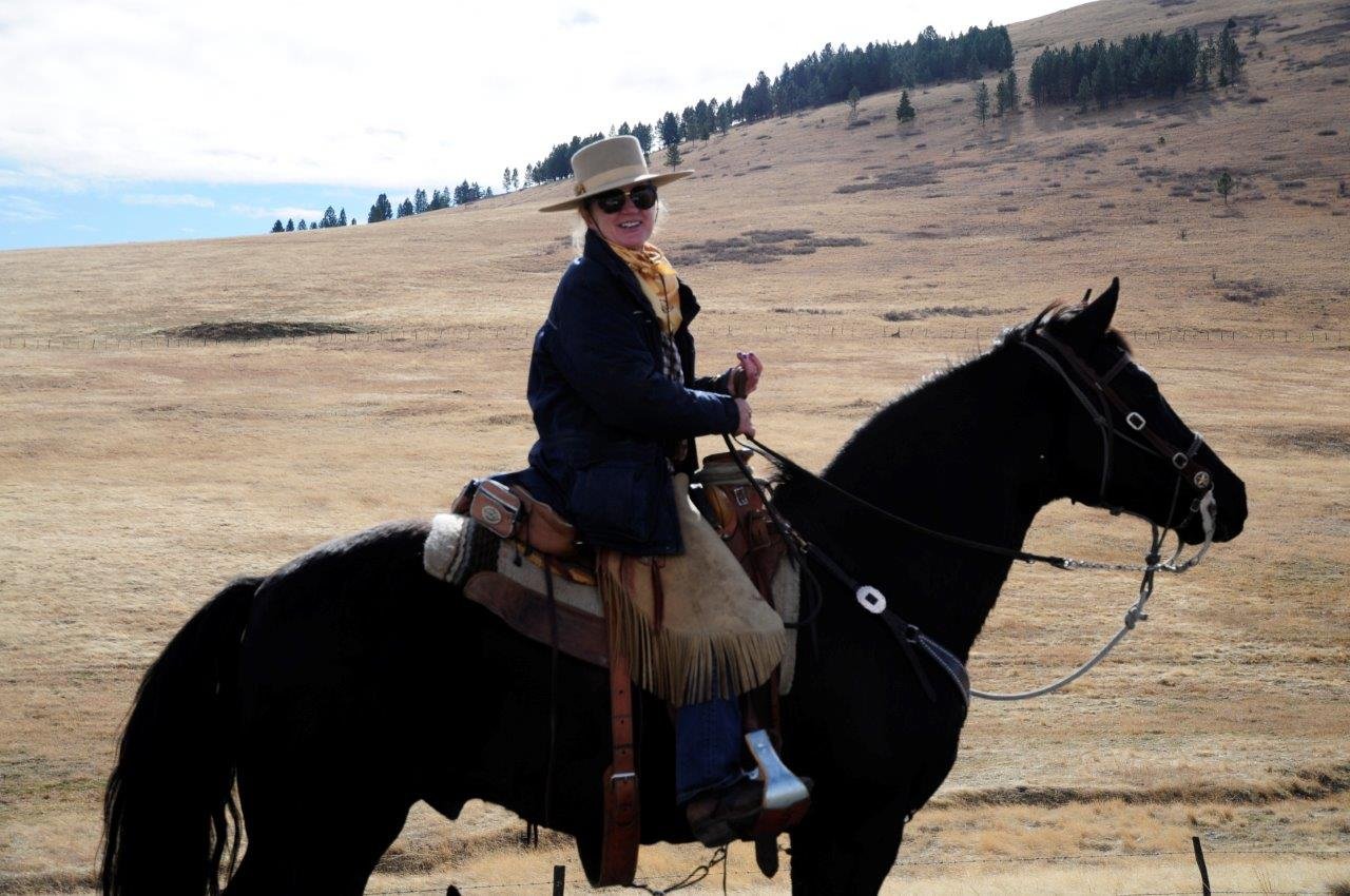 A woman riding a black horse in a dry, open field with rolling hills and sparse trees in the background. She is wearing a wide-brimmed hat, sunglasses, a dark jacket, and a yellow scarf.