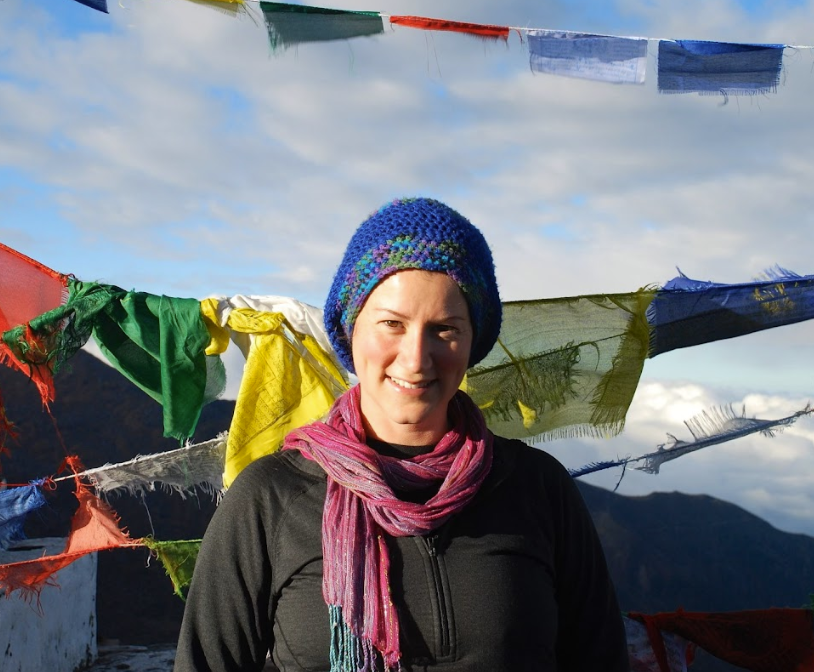 A woman wearing a blue knit hat and pink scarf, smiling outdoors with prayer flags fluttering in the background and mountains under a partly cloudy sky.