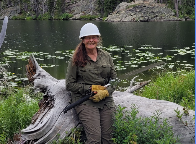 Woman in a white hard hat and yellow work gloves sitting on a fallen tree by a lake, holding a saw, surrounded by green plants and trees.