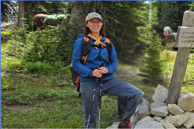 A woman with a backpack standing with one foot on a rock, smiling in a forested area during a hike.