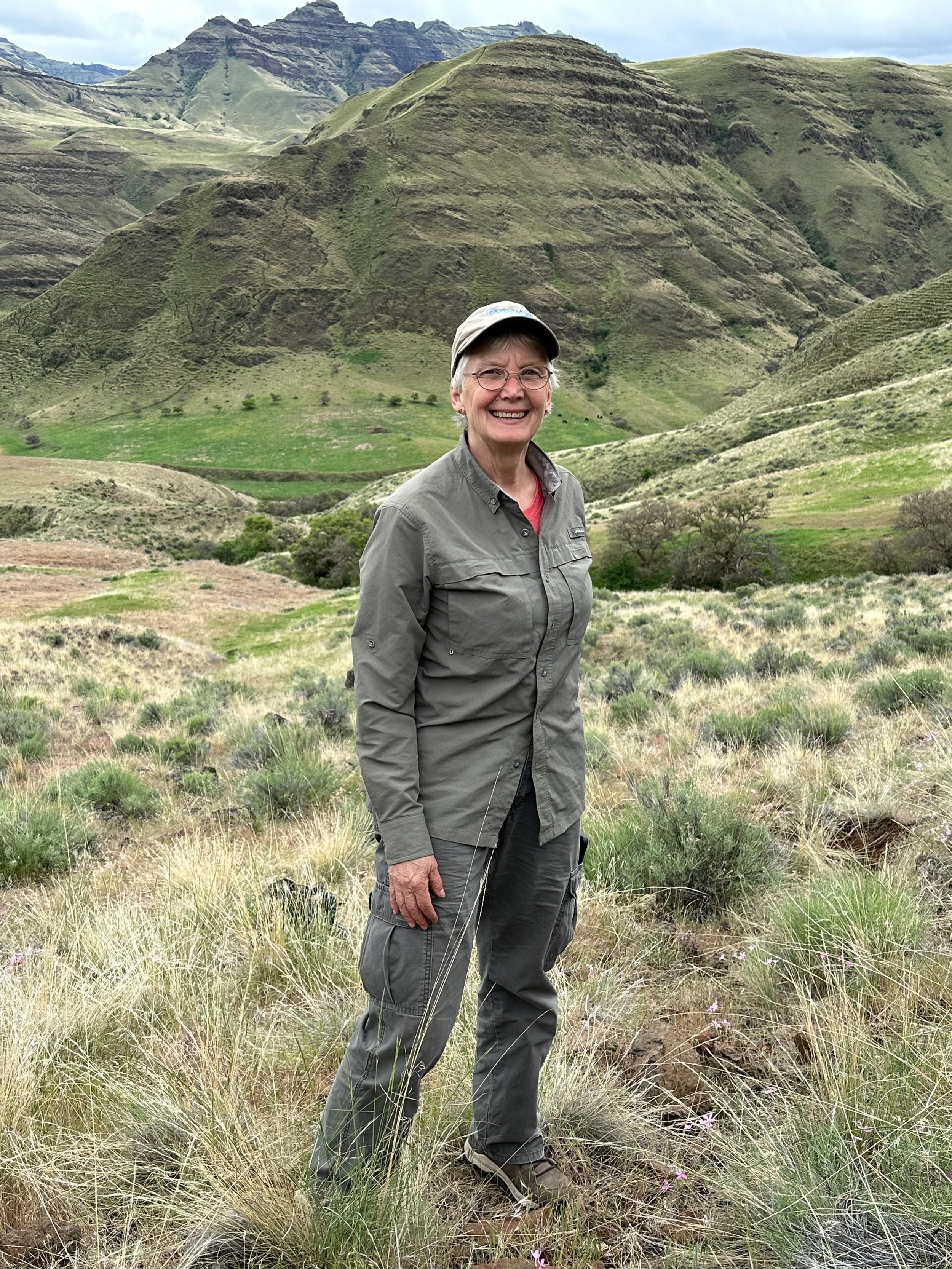 A smiling woman dressed in outdoor hiking attire standing in a green, mountainous landscape with waves of grassy hills and rocky terrain.