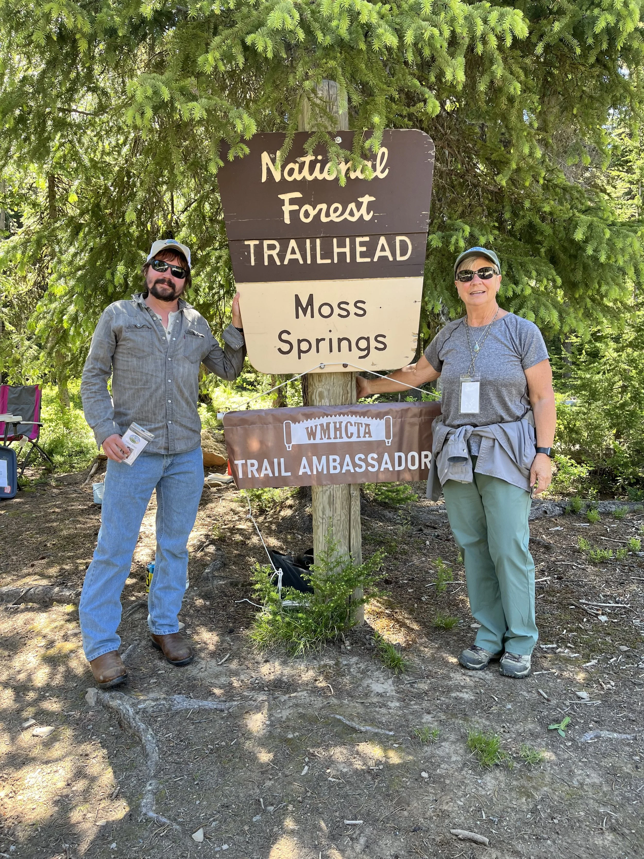 Two people standing next to a sign at the trailhead of a national forest, with trees in the background. The man on the left is wearing sunglasses, a gray shirt, and jeans, and holding a pamphlet. The woman on the right is also wearing sunglasses, a gray t-shirt, and green pants, with a gray jacket tied around her waist. They are holding a rope attached to the sign that reads 'WMMCTA Trail Ambassador.'