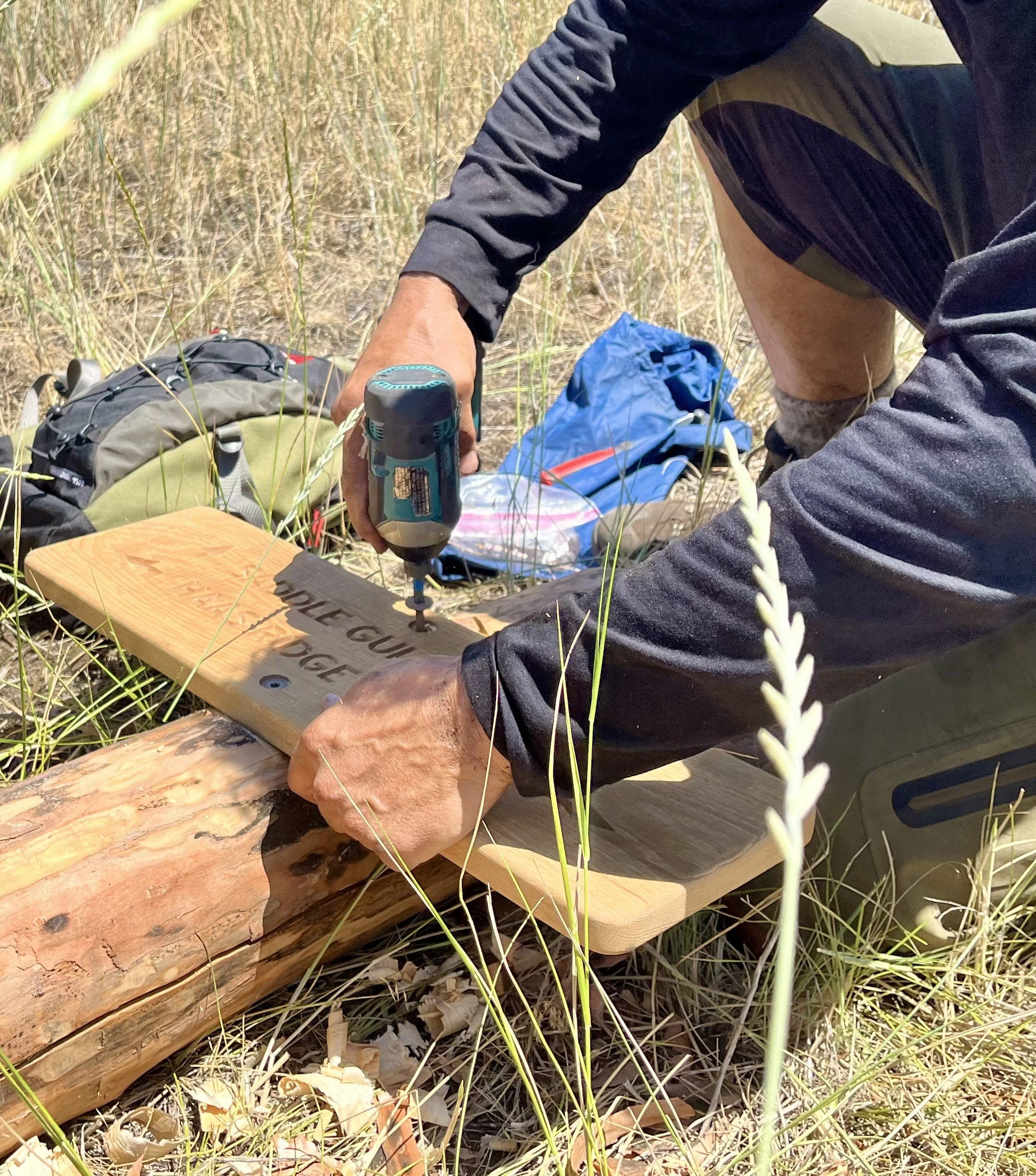 A person using a cordless drill to write on a wooden board in a grassy outdoor setting, with backpacks and supplies nearby.